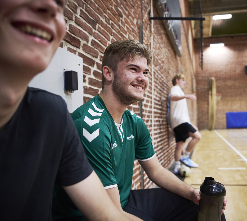 Smilende elever sidder på sidelinjen til idræt